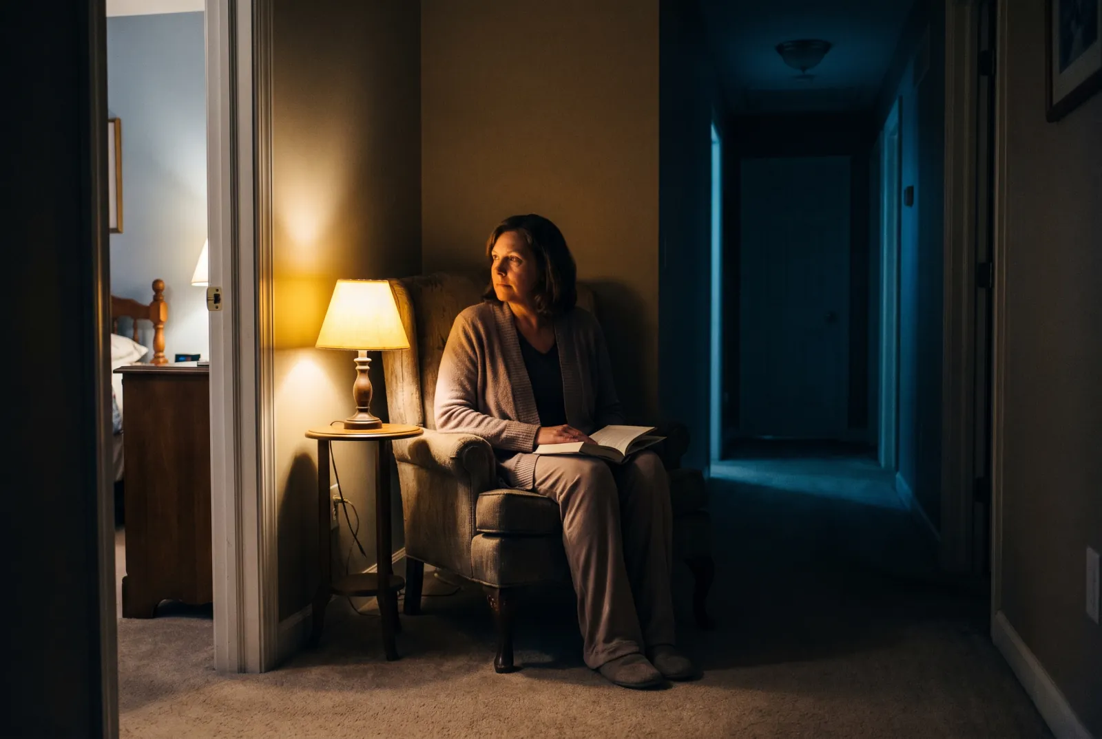 A caregiver sitting awake with a book in a lamplit chair outside a bedroom door.